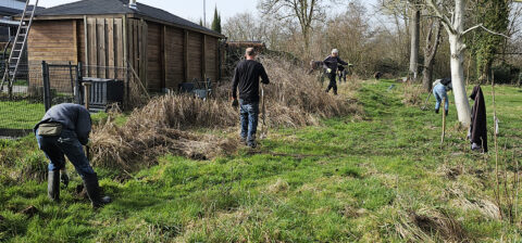 Mensen wieden gras en planten bomen naast een houten schuur in een groen, bosrijk gebied.