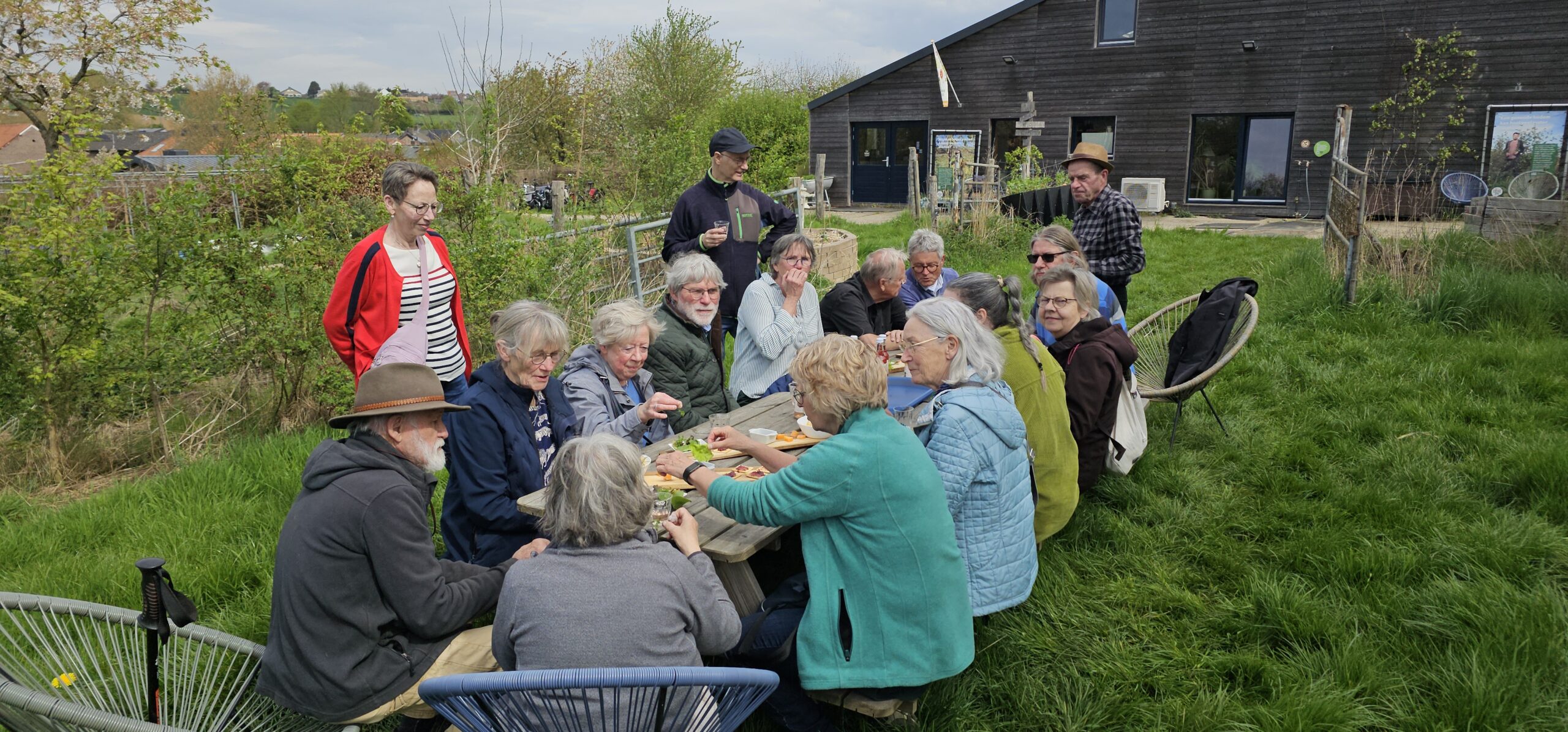 Groep mensen zit buiten aan een picknicktafel in een tuin, omgeven door bomen en een gebouw.