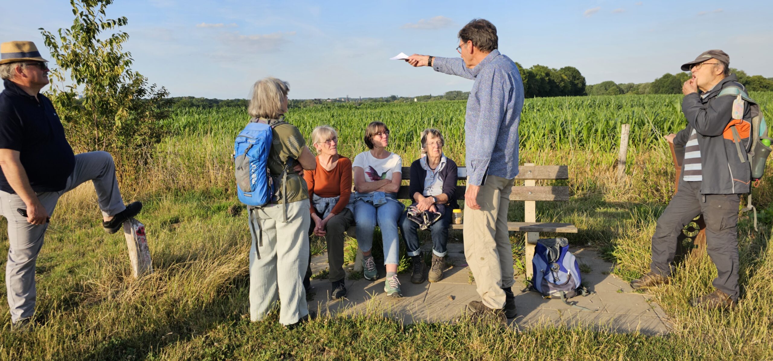 Groep mensen in gesprek op een bankje langs een pad in een groen landschap.