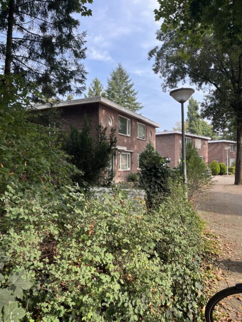 Bakstenen huis omringd door groen, met lantaarnpaal en fietswiel zichtbaar op zonnige dag.
