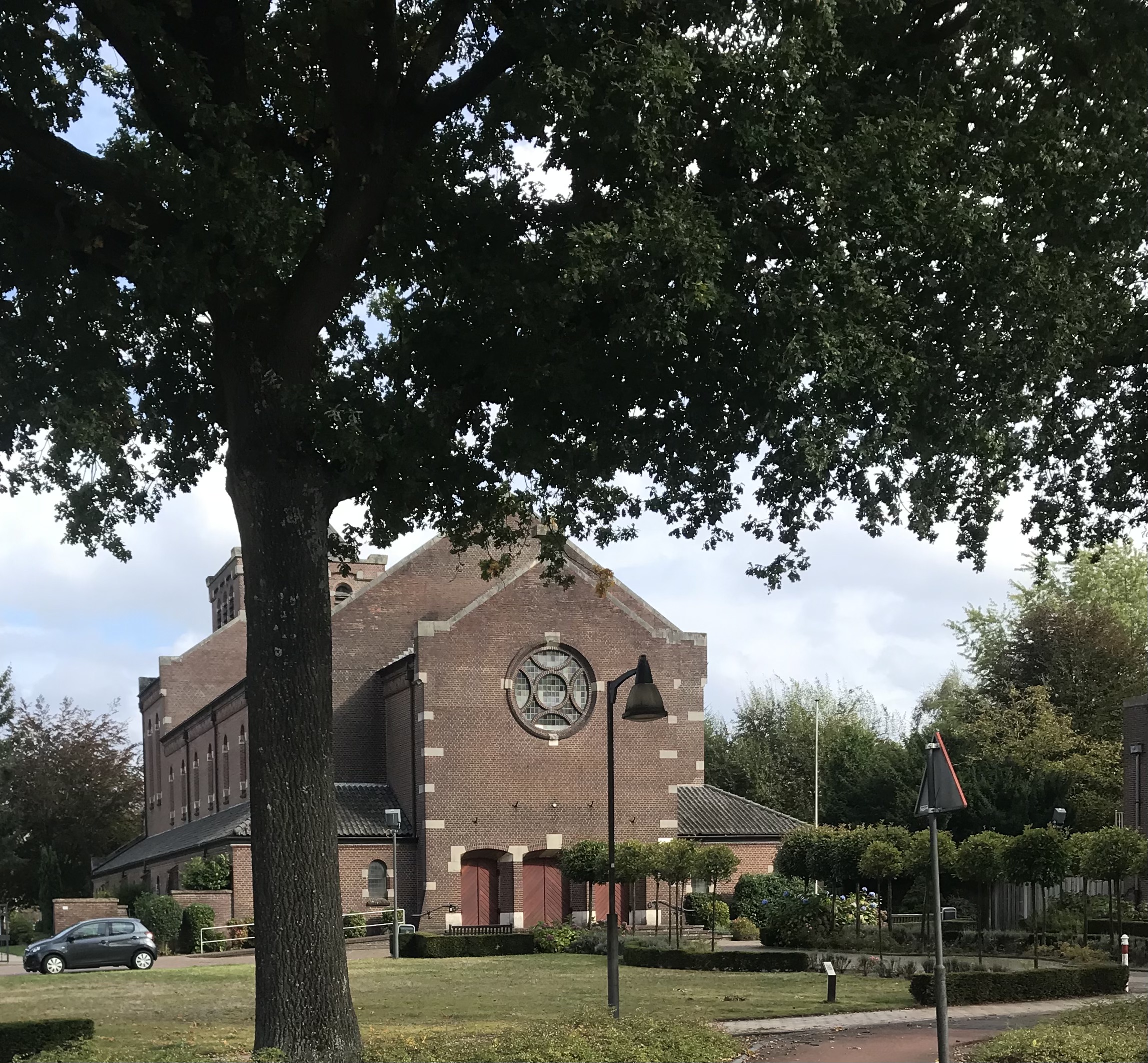Bakstenen kerkgebouw met rozetraam, omgeven door bomen en een grasveld; lantaarnpaal op de voorgrond.