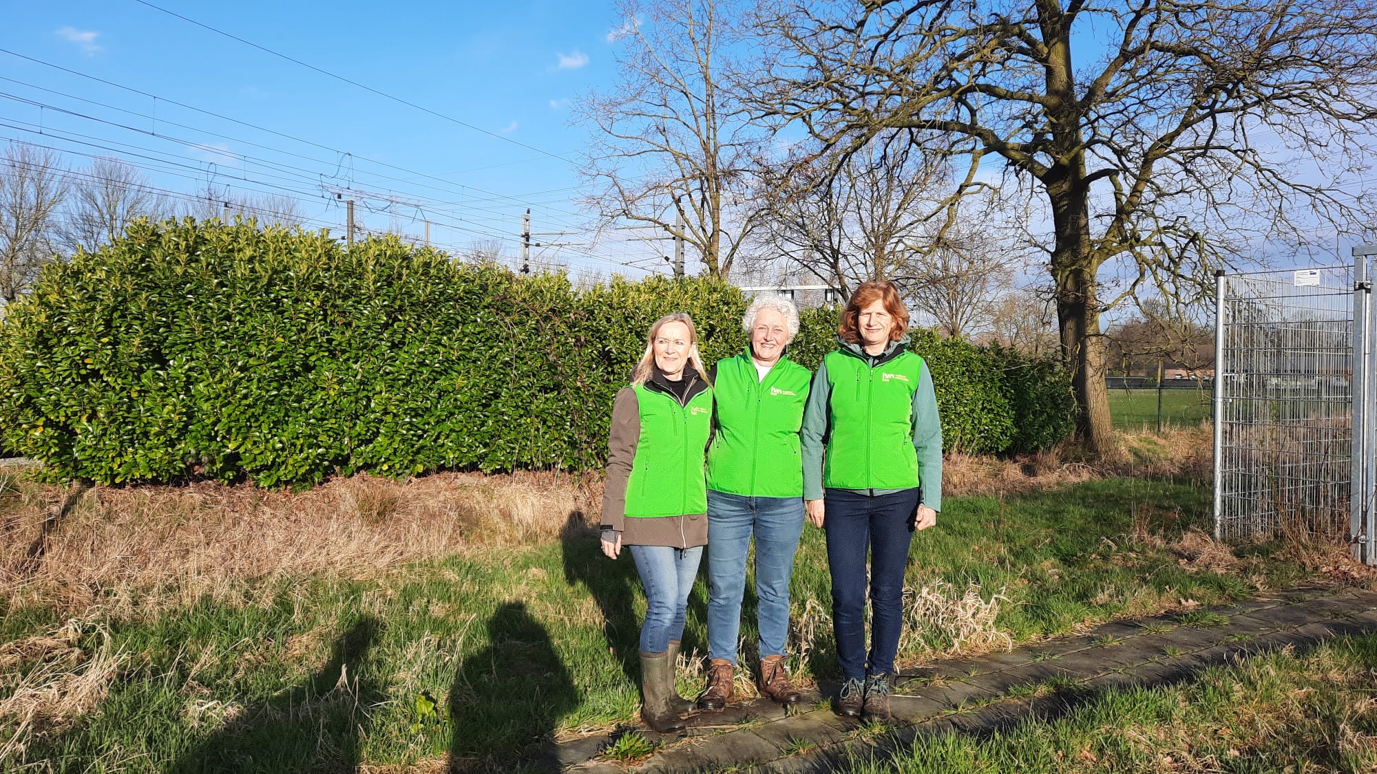 Drie mensen in groene jassen staan buiten naast een heg en een boom, met blauwe lucht.