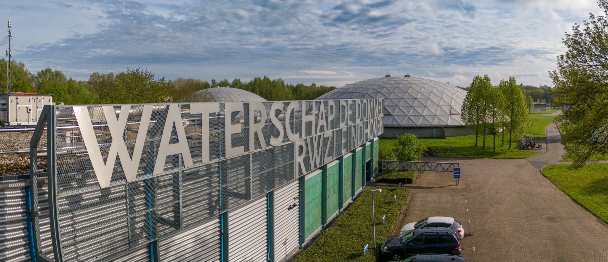Gebouw met "Waterschap De Dommel" en koepelvormige structuur op zonnige dag, omgeven door groen.