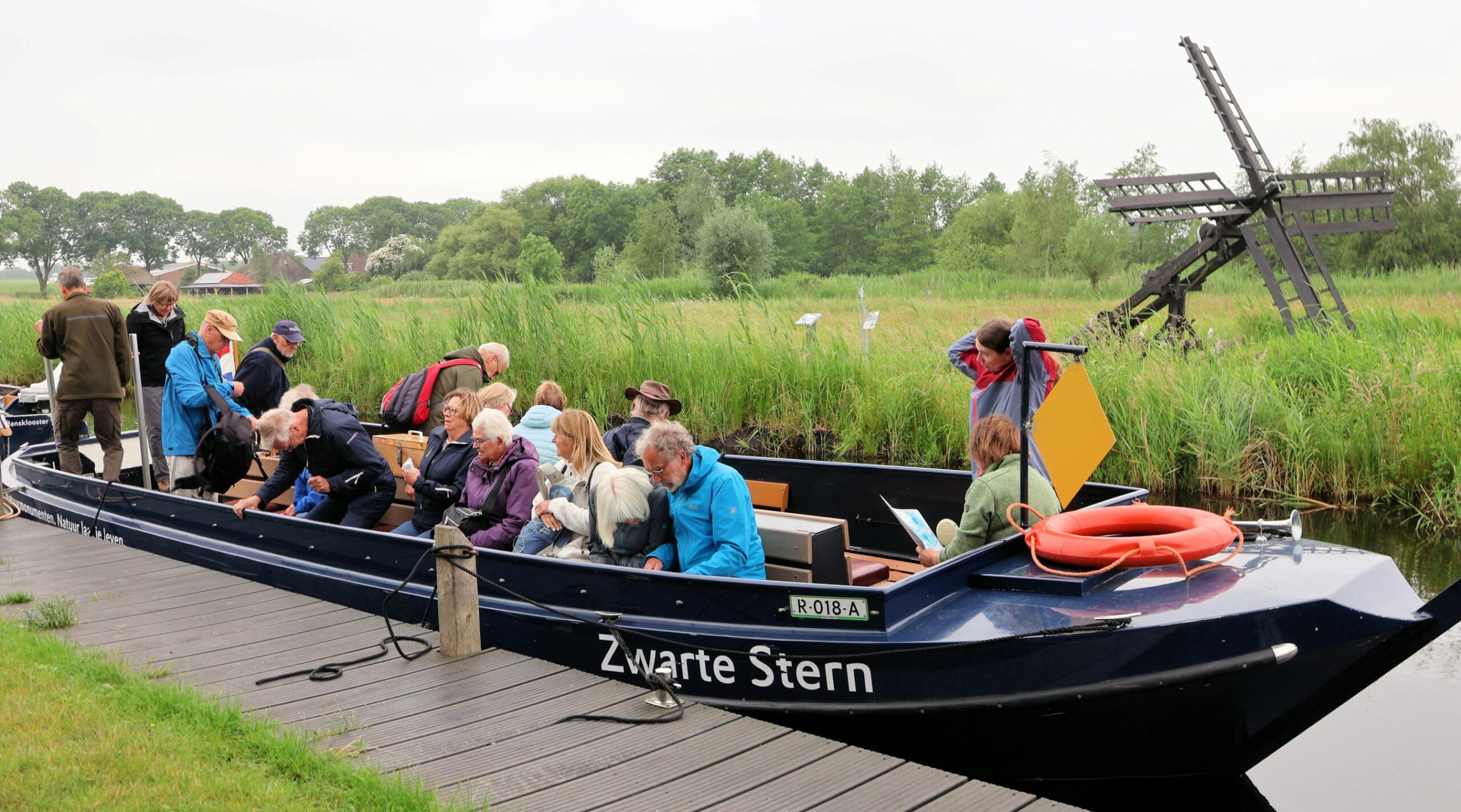 Groep mensen in een boot op kanaal, met molen en riet op achtergrond. Plaatsnaam "Zwarte Stern".