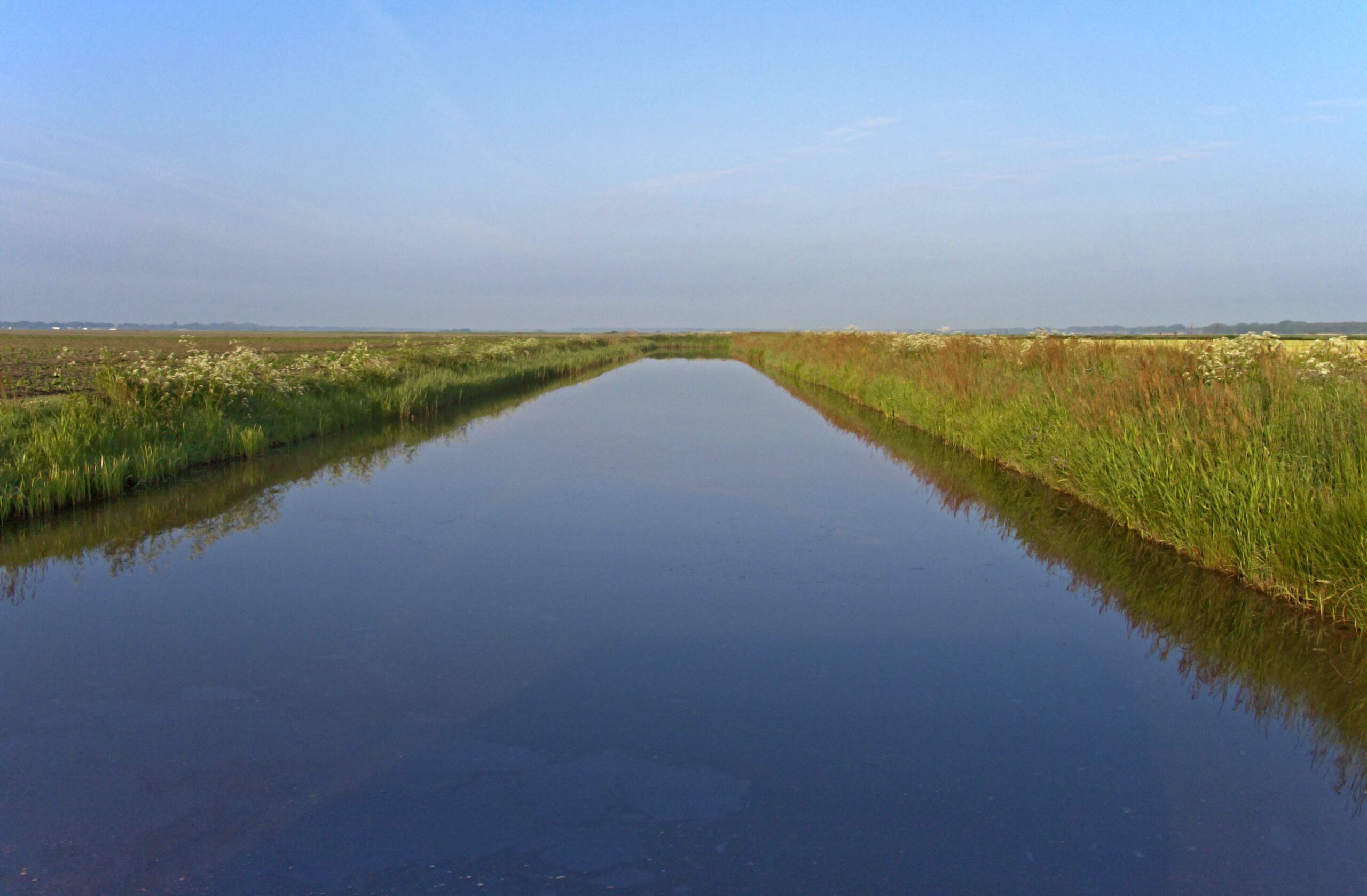 Lang kanaal omringd door groene velden en blauwe lucht.