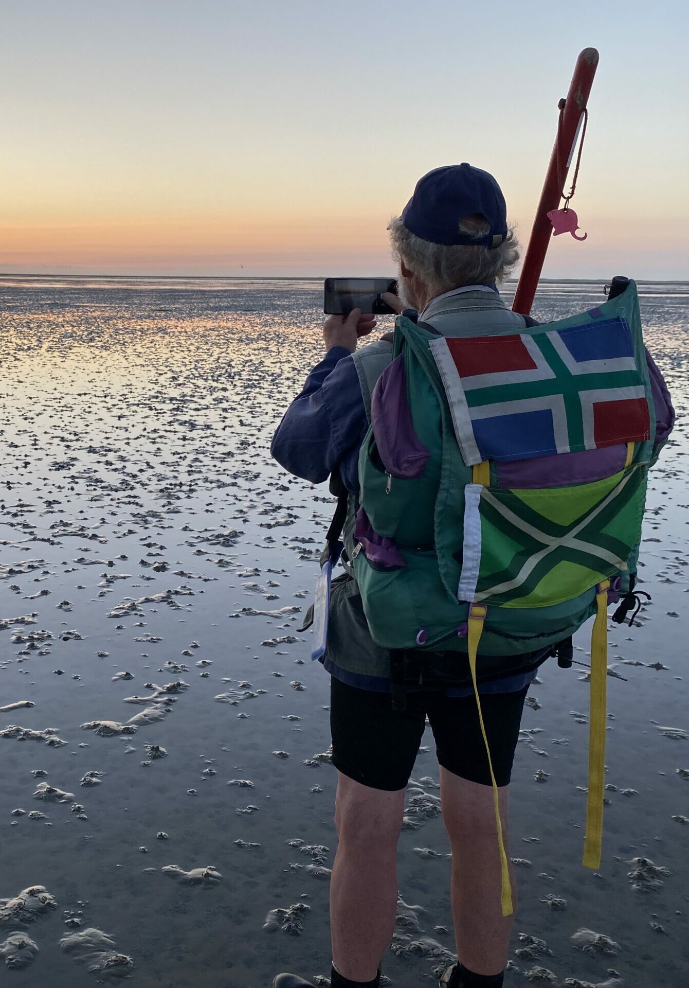 Persoon met kleurrijke rugzak fotografeert zonsopgang op een moddervlakte bij laagwater.