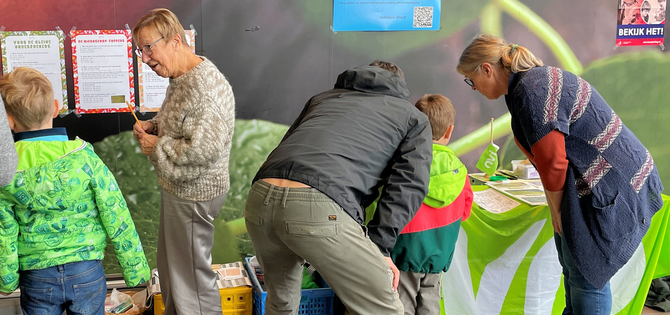 Mensen buigen zich over een tafel met activiteiten, omgeven door kleurrijke displays en documenten.