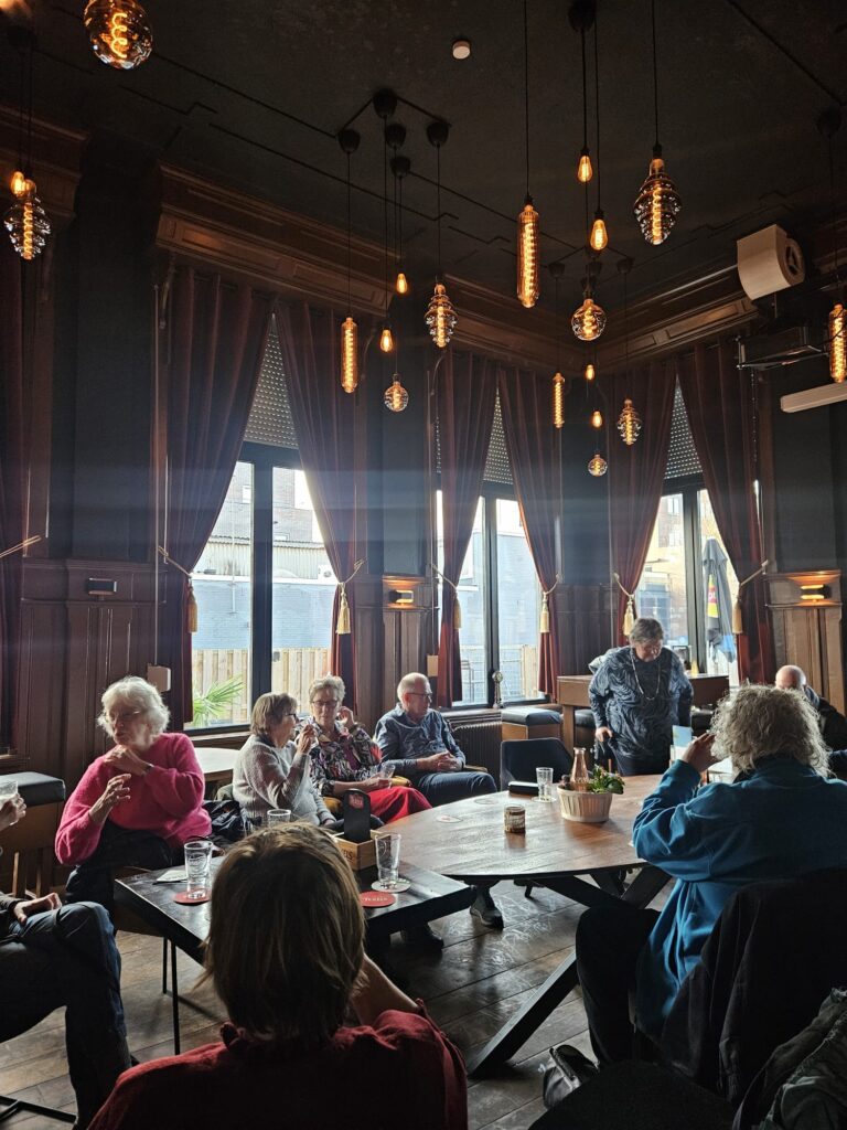 Groep mensen in café-achtige ruimte met hoge plafonds en hanglampen.