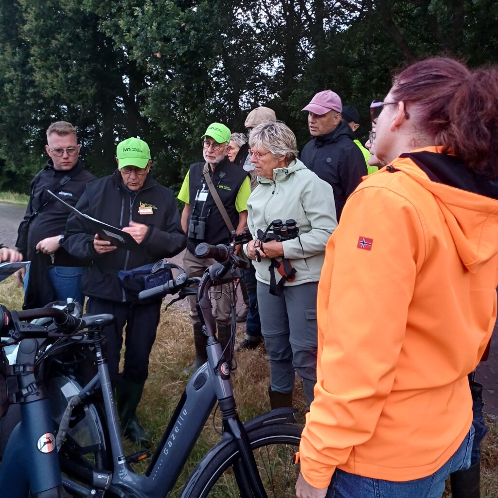 Groep mensen met fietsen, enkele met verrekijkers, in een bosrijke omgeving.
