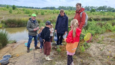 Groep mensen bij een vijver in de natuur, kinderen met schepnetten, groene omgeving.