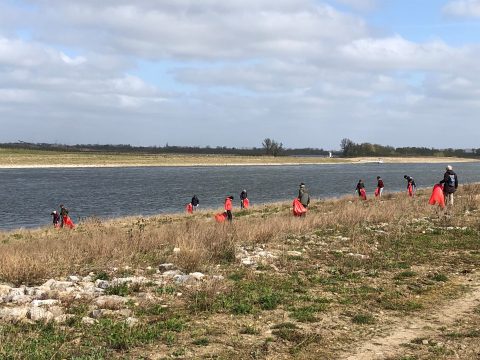 Mensen ruimen afval langs een rivier op, met rode vuilniszakken in een natuurlijke omgeving.
