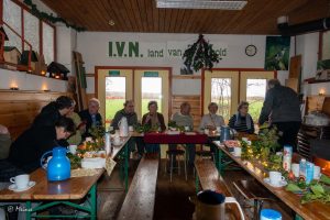 Groep mensen aan tafel in knusse ruimte met IVN-bord, kerstdecoraties en drankjes.