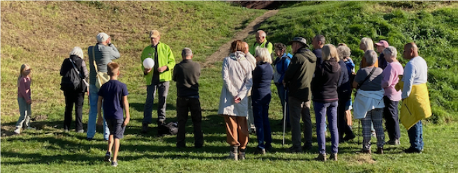 Groep mensen verzameld in een park, luisterend naar een man met een groen jack en een witte bal.