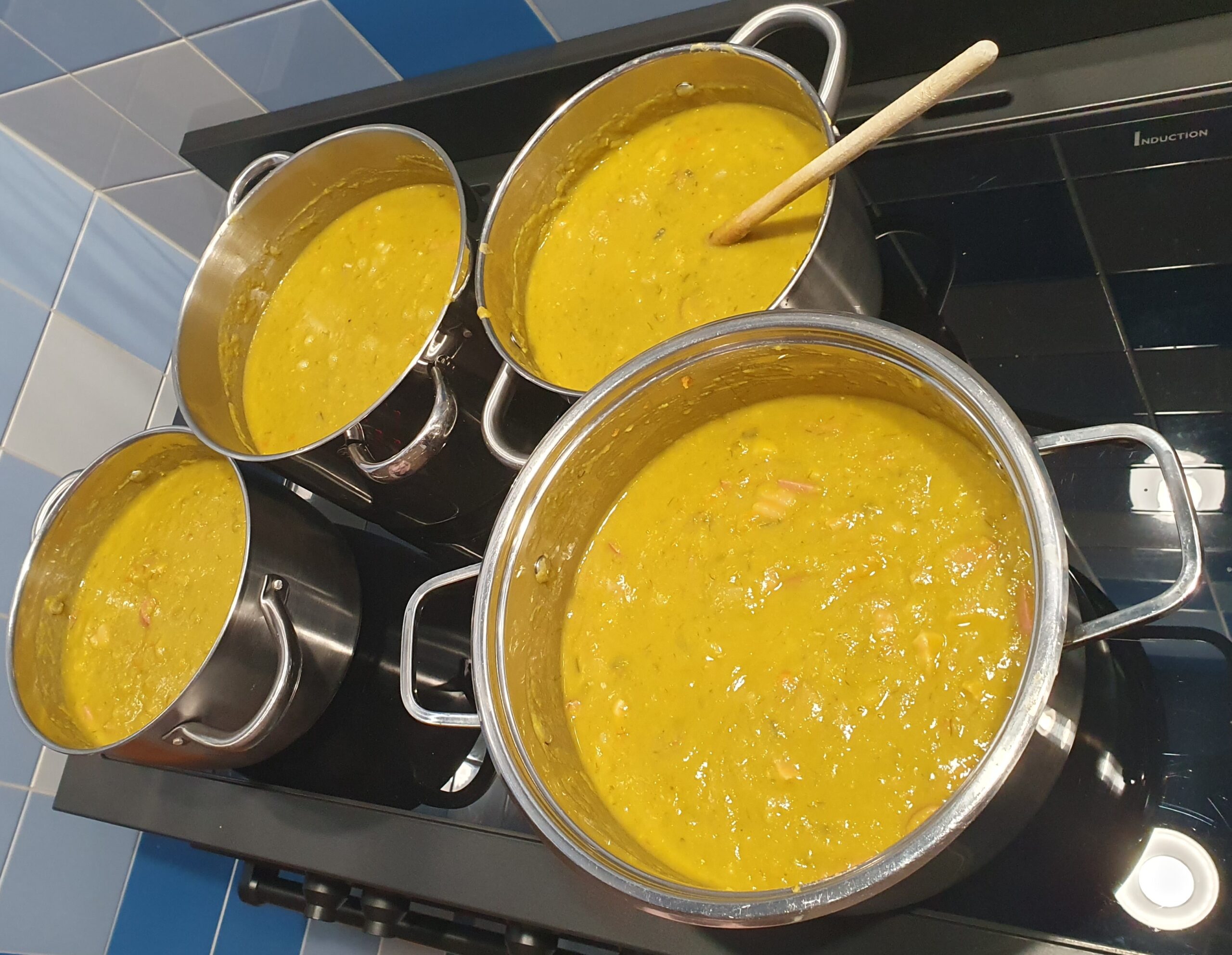 Vier grote pannen met erwtensoep op een kookplaat, één met een houten lepel erin.
