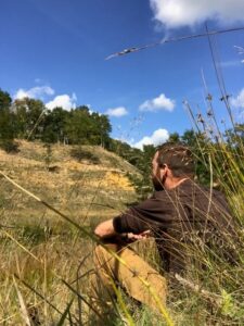Man zit in gras, kijkt uit over heuvelachtig landschap met blauwe lucht en wolken.