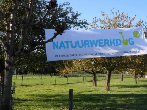 Spandoek met tekst "Natuurwerkdag" hangt tussen bomen in een groene omgeving.