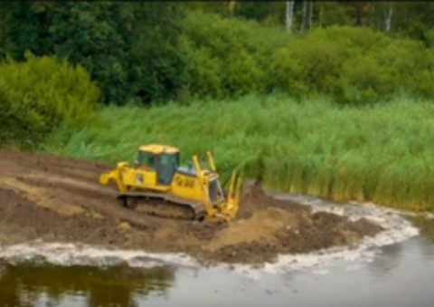 Een gele bulldozer verplaatst aarde langs een groen rietland bij een waterkant.