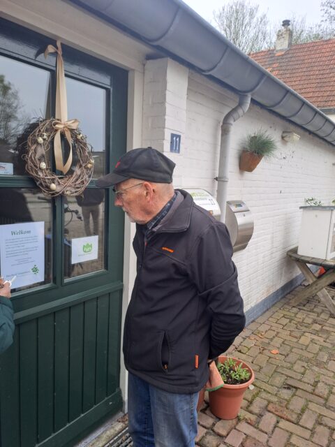 Man leest een brief bij een groene deur van huis nummer 11 met krans en planten langs de muur.