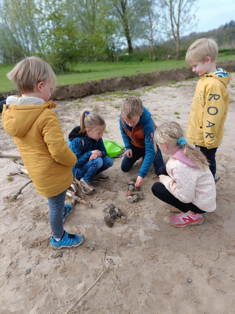 Kinderen verzamelen stenen op een zandgrond, met gras en bomen op de achtergrond.