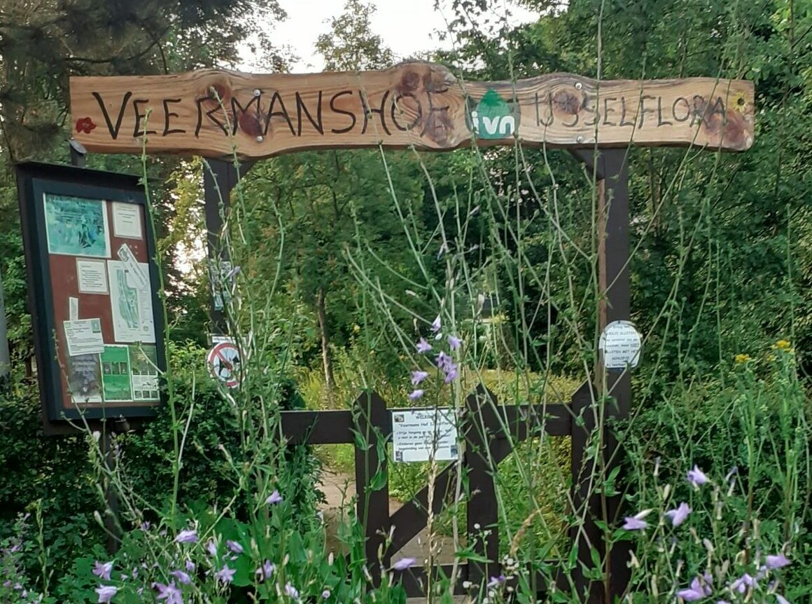 Houten toegangspoort van Veerenshof met bloemen en infobord, omgeven door groene bomen.