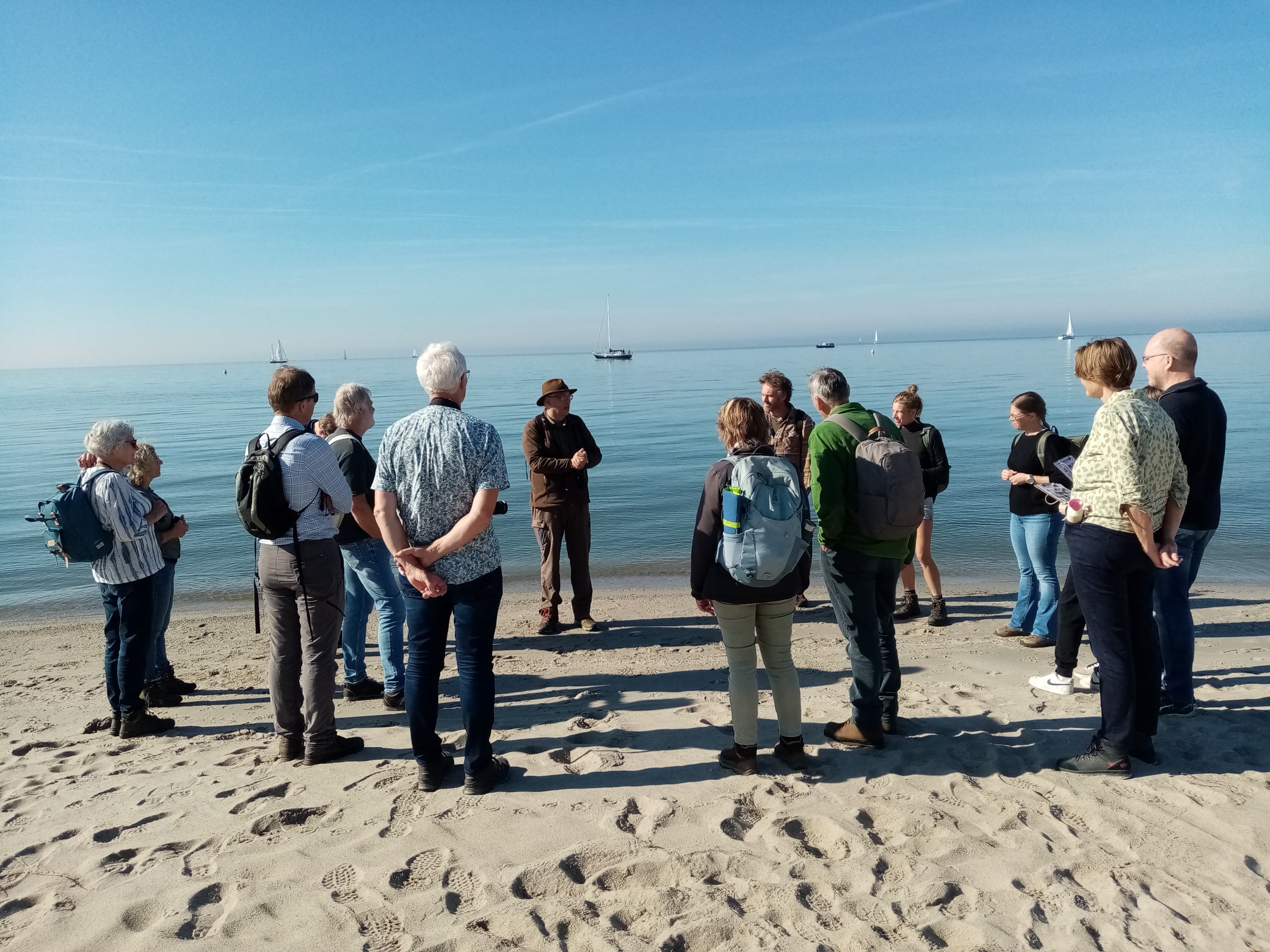 Een groep mensen luistert naar een spreker op een zandstrand met rustige zee op de achtergrond.