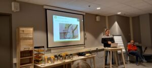 Man presenteert voor een projectiescherm met uilenafbeelding, omgeven door stoelen en koffieapparatuur.