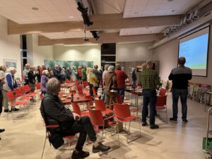 Groep mensen kijkt naar een presentatie in een zaal met rode stoelen en kunstwerken aan de muur.
