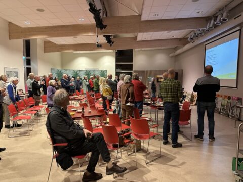 Groep mensen kijkt naar een presentatie in een zaal met rode stoelen en kunstwerken aan de muur.