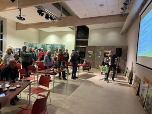 Een groep mensen kijkt naar een presentatie in een zaal; een muzikant speelt accordeon op het podium.
