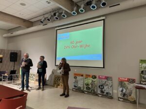Mensen op podium vieren 60 jaar IVN Olst-Wijhe met presentatie en natuurposters op achtergrond.