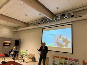 Een man presenteert in een zaal met een projectiescherm en natuurposters, terwijl een ander aan een tafel zit.