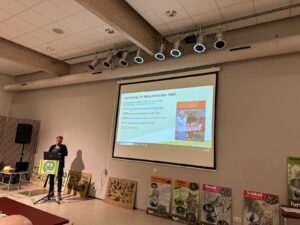 Persoon presenteert naast projectiescherm met tekst over "Natuurvrienden 1965" en vogelinformatiepanelen.