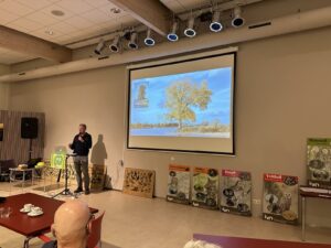 Man geeft presentatie met boom op scherm; natuuraffiches op de achtergrond.