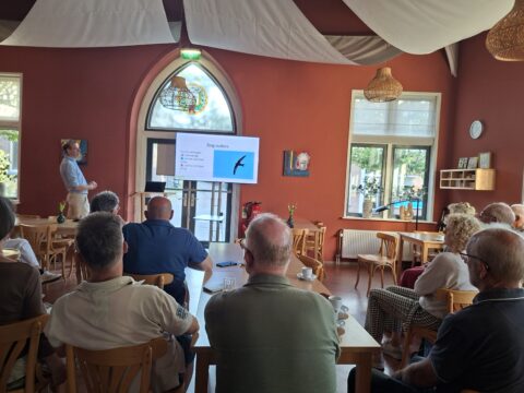 Persoon presenteert voor een groep in een zaal met scherm en stoelen, warme verlichting.