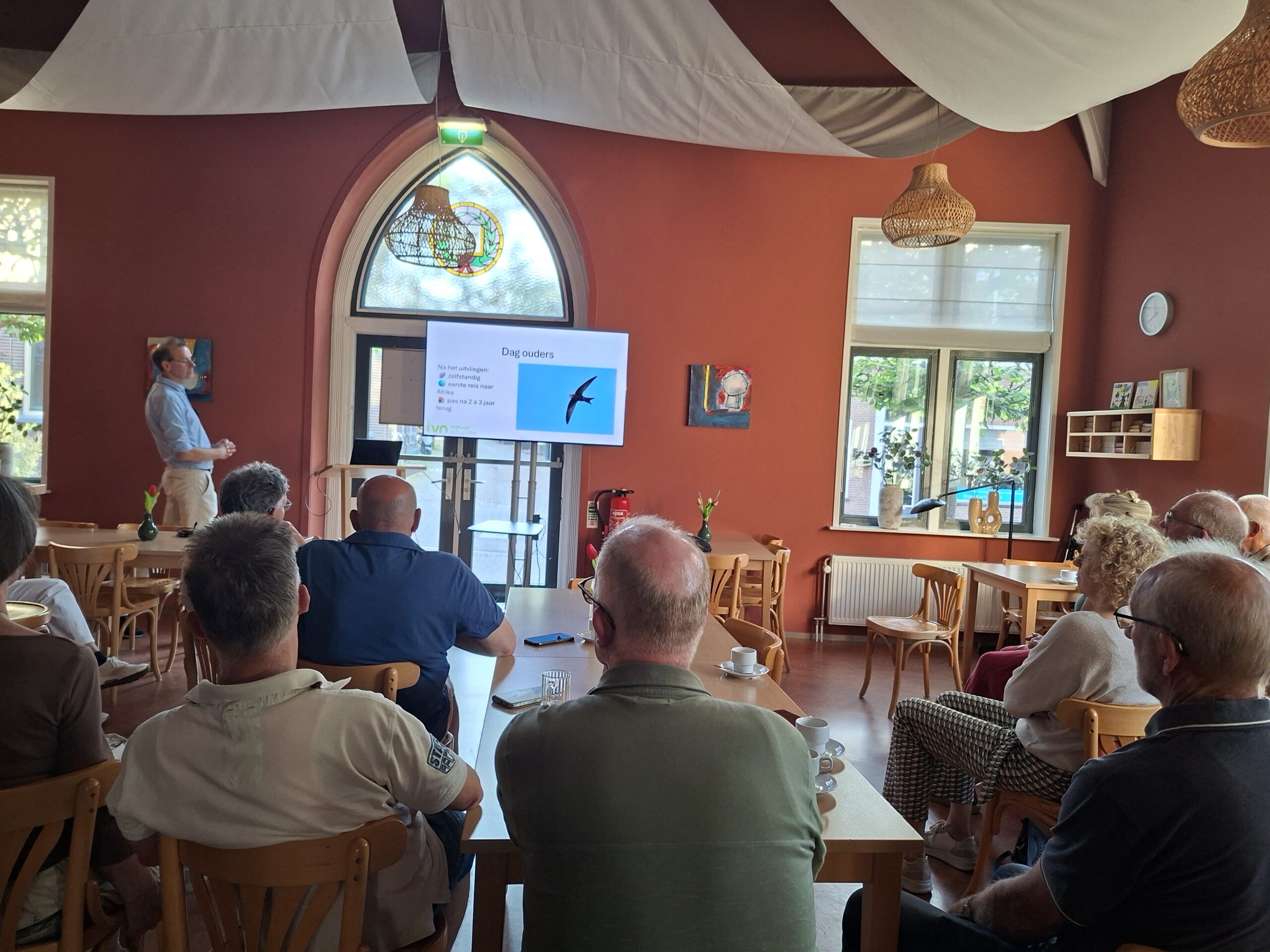 Persoon presenteert voor een groep in een zaal met scherm en stoelen, warme verlichting.