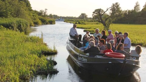 Groep mensen op een boottocht door smalle rivier met gras en bomen langs de oevers.