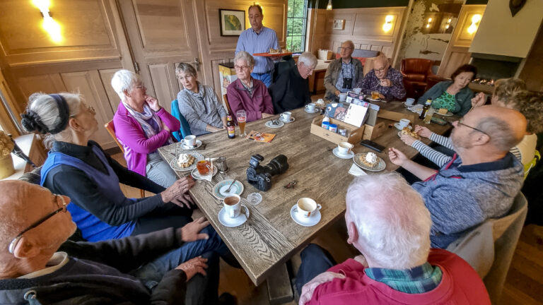 Groep oudere mensen zit aan tafel met koffie en gebak in een gezellig café.