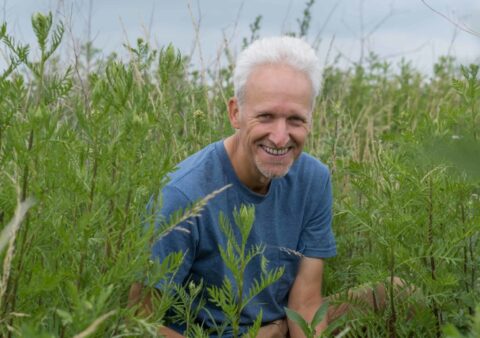 Man zit glimlachend in hoog gras, blauwe lucht op achtergrond.