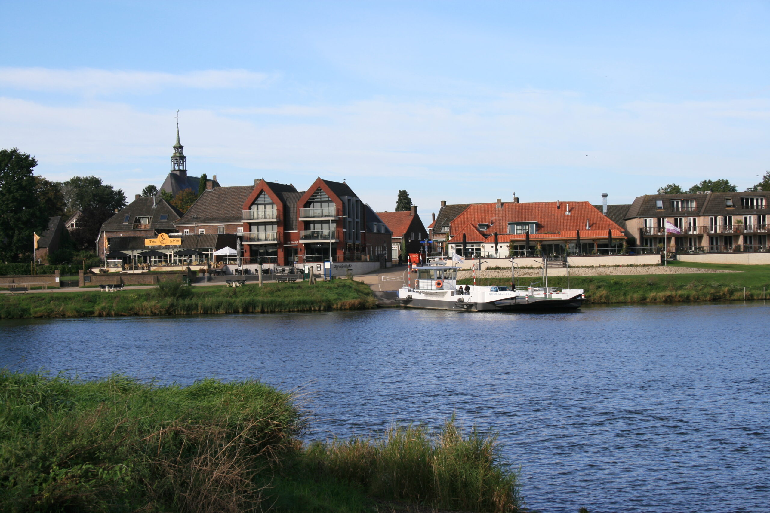 Veerboot aan de rivier voor een dorp met huizen en een kerktoren, omringd door groen.