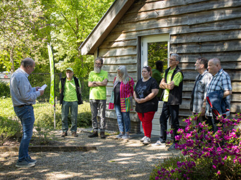 Groep mensen luistert naar spreker buiten naast houten gebouw, omringd door groen.