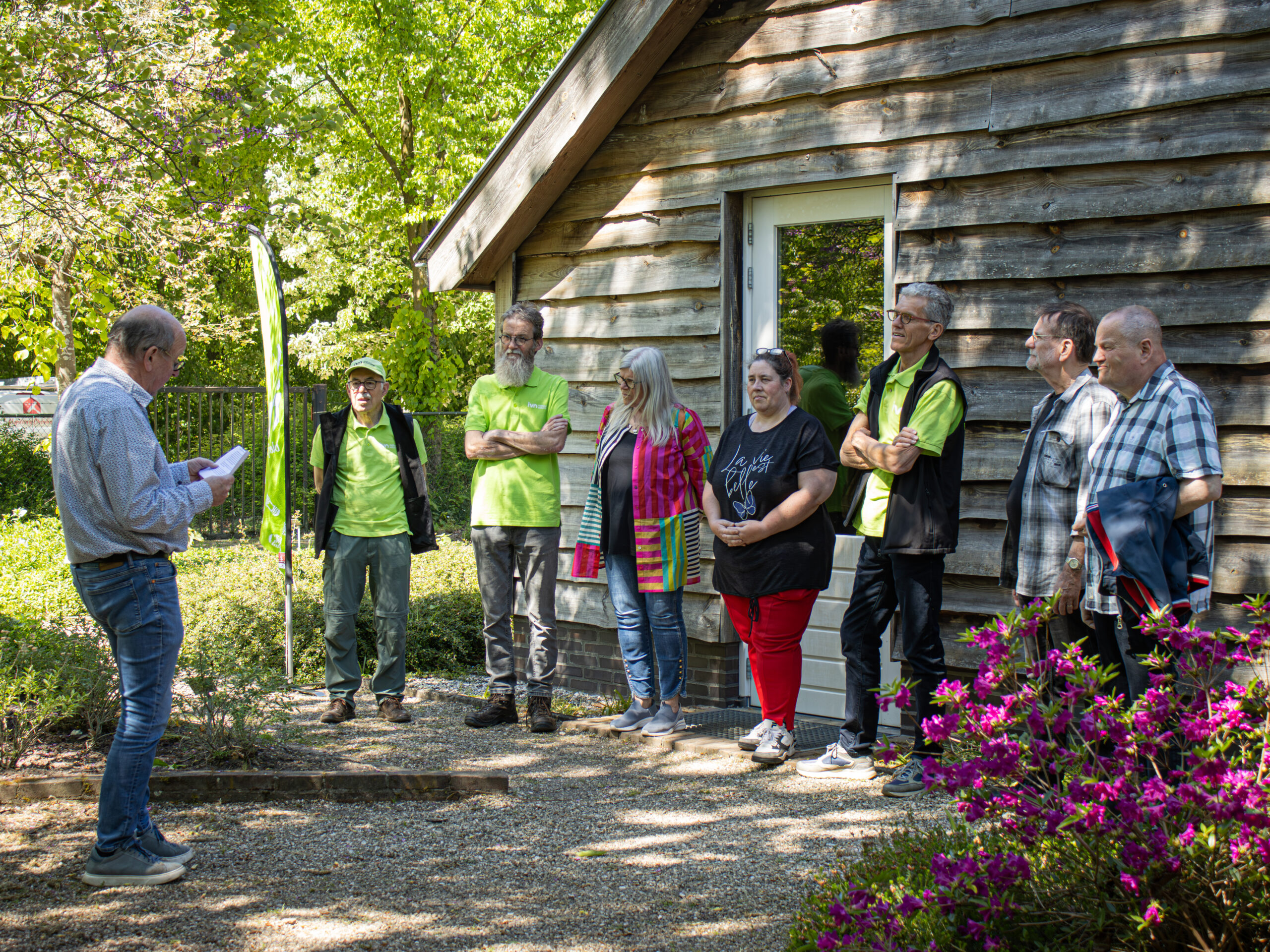 Groep mensen luistert naar spreker buiten naast houten gebouw, omringd door groen.