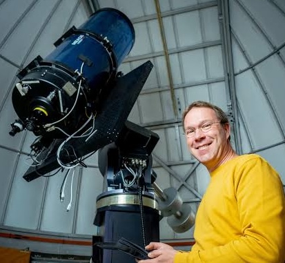 Man in gele trui bij telescoop in observatorium. Lachend naast groot wetenschappelijk instrument.