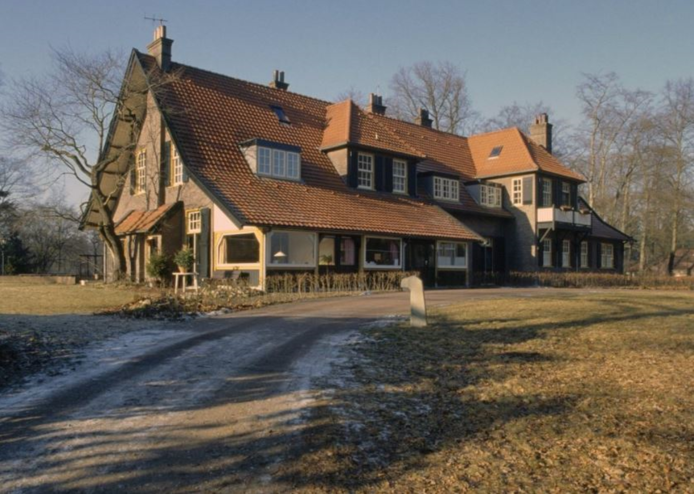 Groot huis met rood pannendak, oprijlaan en kale bomen in een winterse omgeving.
