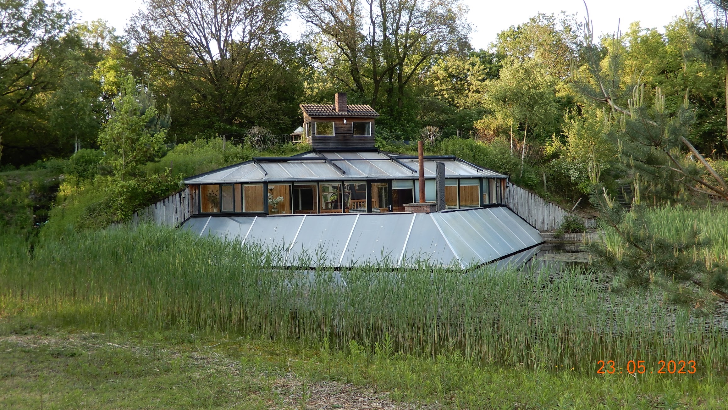 Aardhuis deels ondergronds, omringd door groen, met glazen dak en houten bovenbouw.