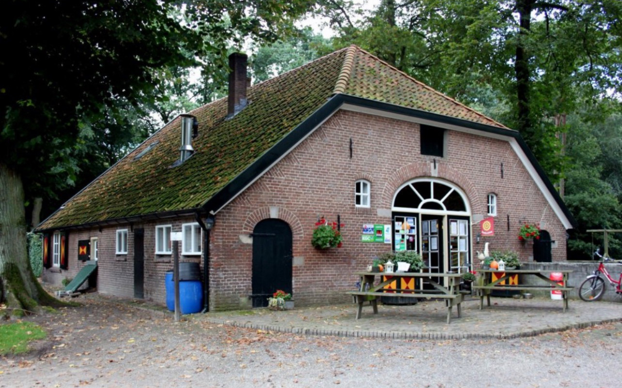 Bakstenen boerderij met groene deur, houten picknicktafels, en fietsen; omringd door bomen.