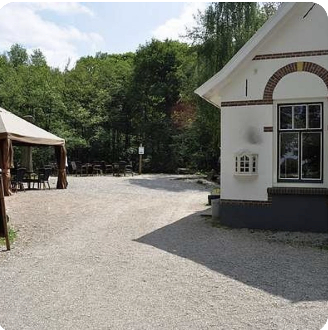 Gebouw met raam rechts en tent met stoelen links, omgeven door bomen en grindpad.