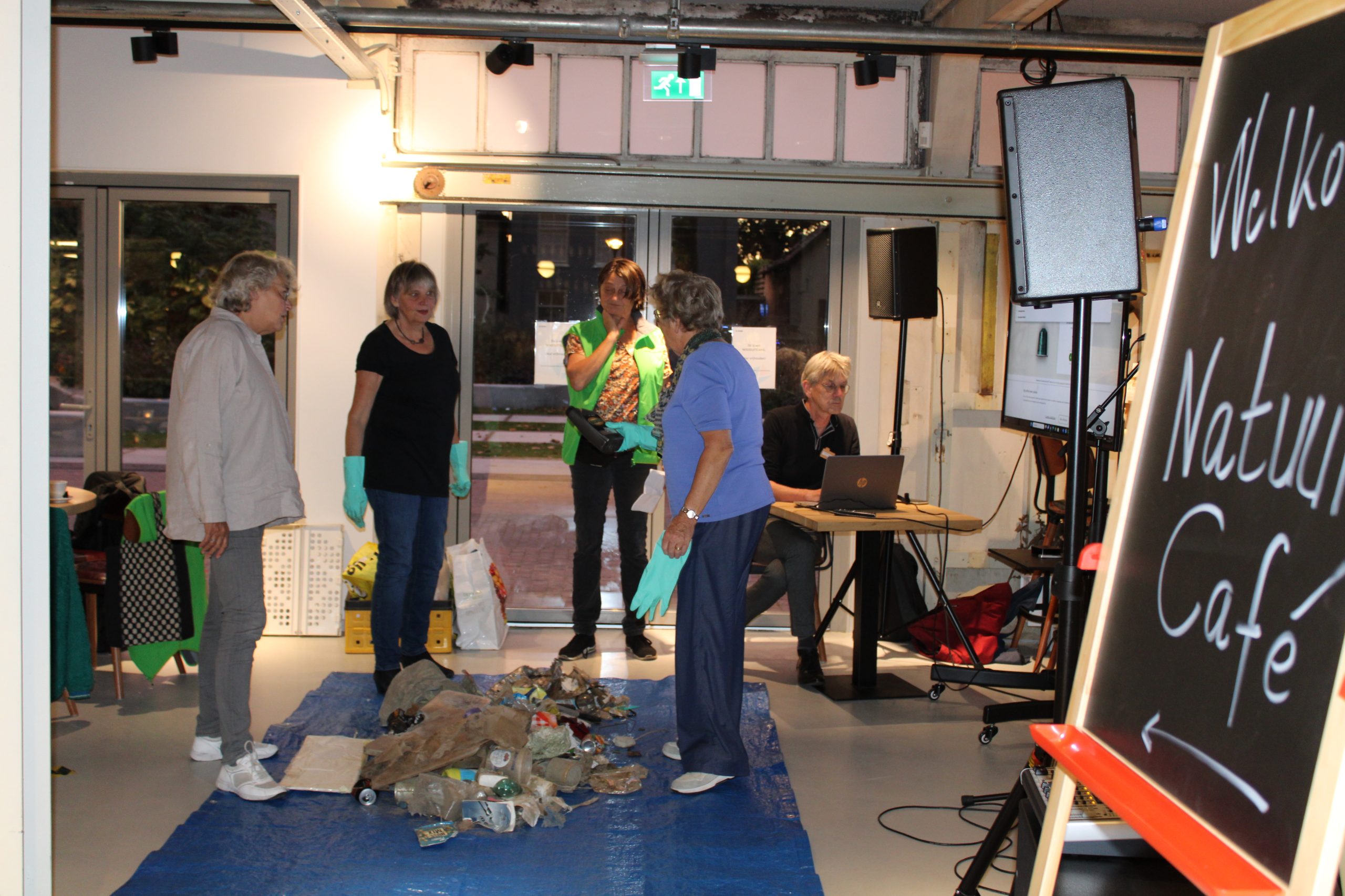 Mensen sorteren afval op een blauwe doek in een zaal met een bord "Natuur Café".