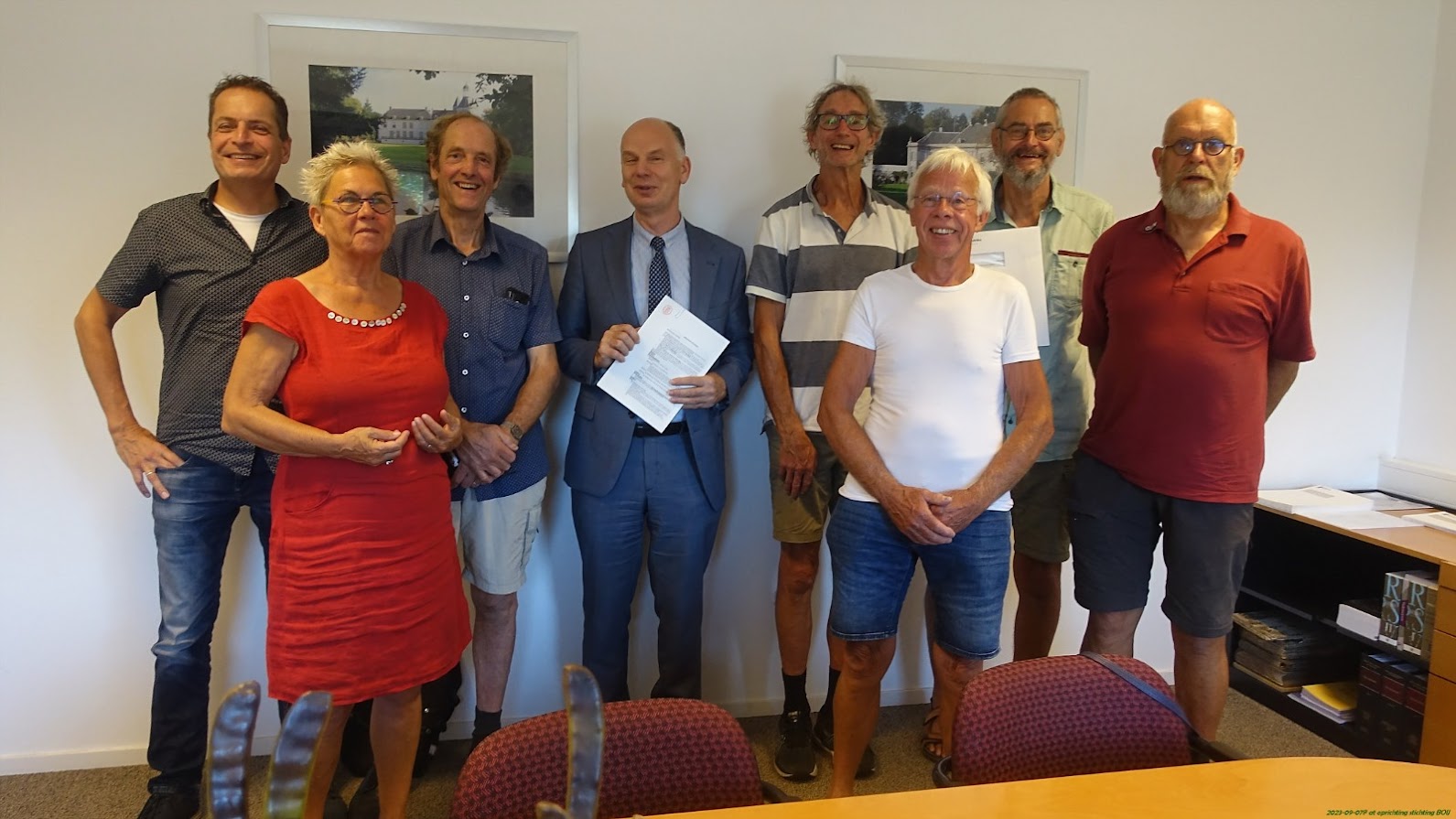 Groep van acht mensen poseert in een kantoor, een man in pak houdt een document vast.