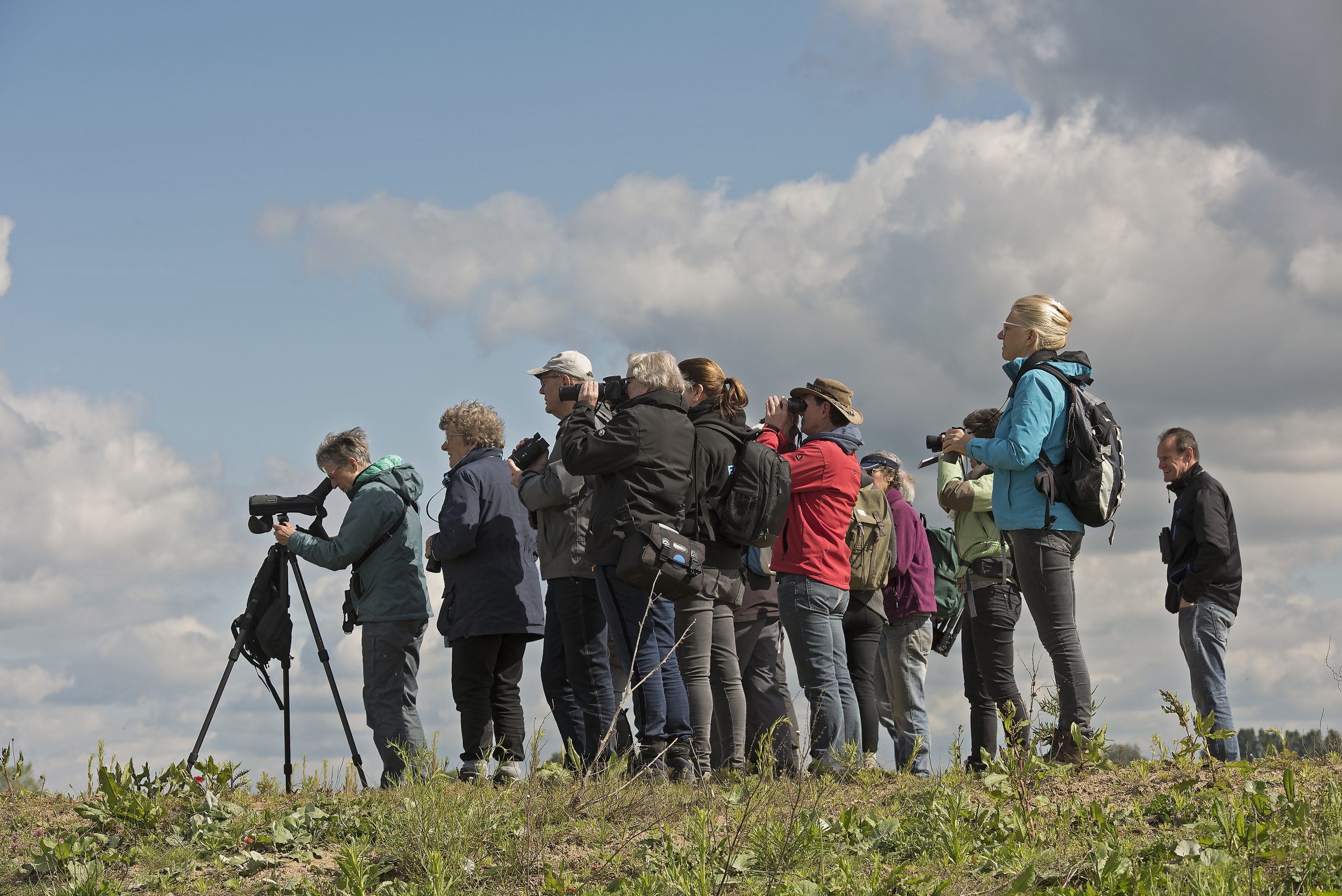 Groep mensen met camera's en verrekijkers kijkt naar de lucht op een grasheuvel onder bewolkte hemel.