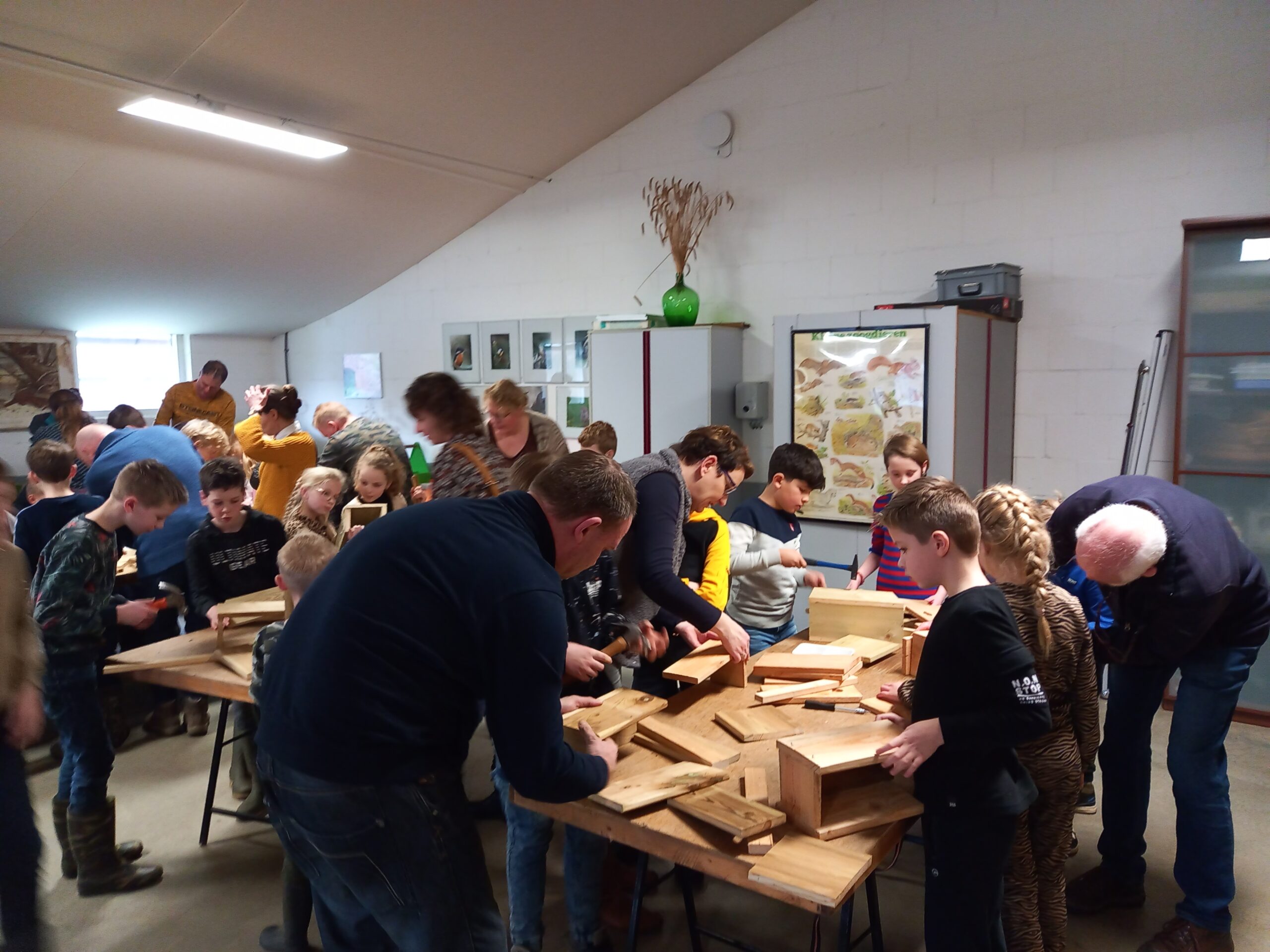 Groep kinderen en volwassenen maakt houten vogelhuisjes in een klaslokaal.
